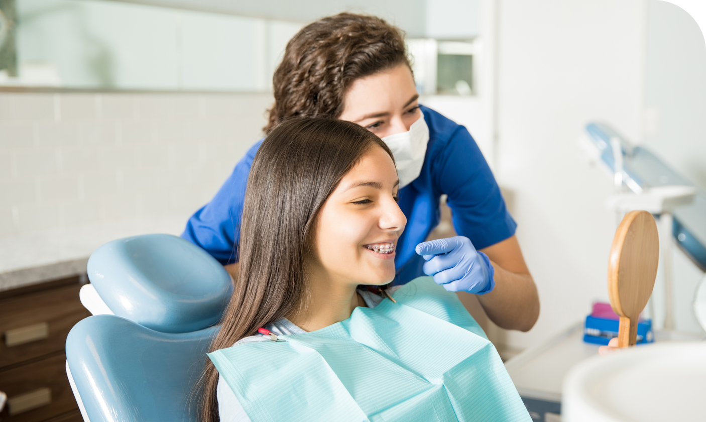 Young girl with braces smiling at her reflection while a dental hygienist checks her teeth in a clinic.